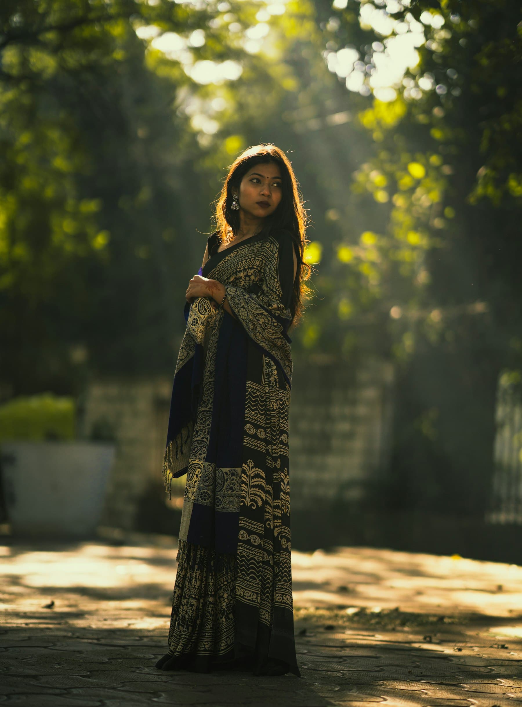 A woman in a dark printed saree stands in morning light under trees, looking over her shoulder