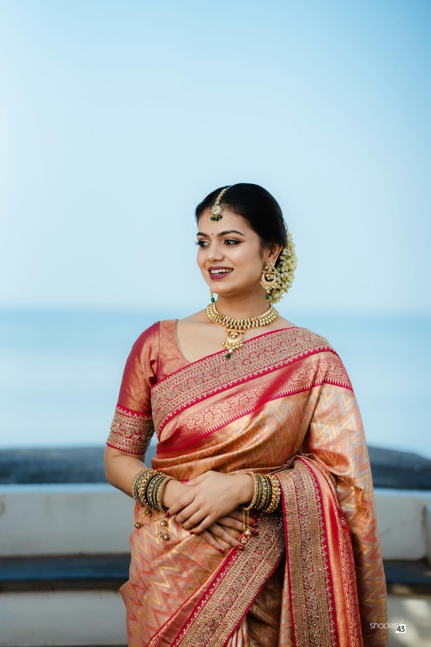 A woman in a peach and red silk saree smiles against a clear sky by the sea