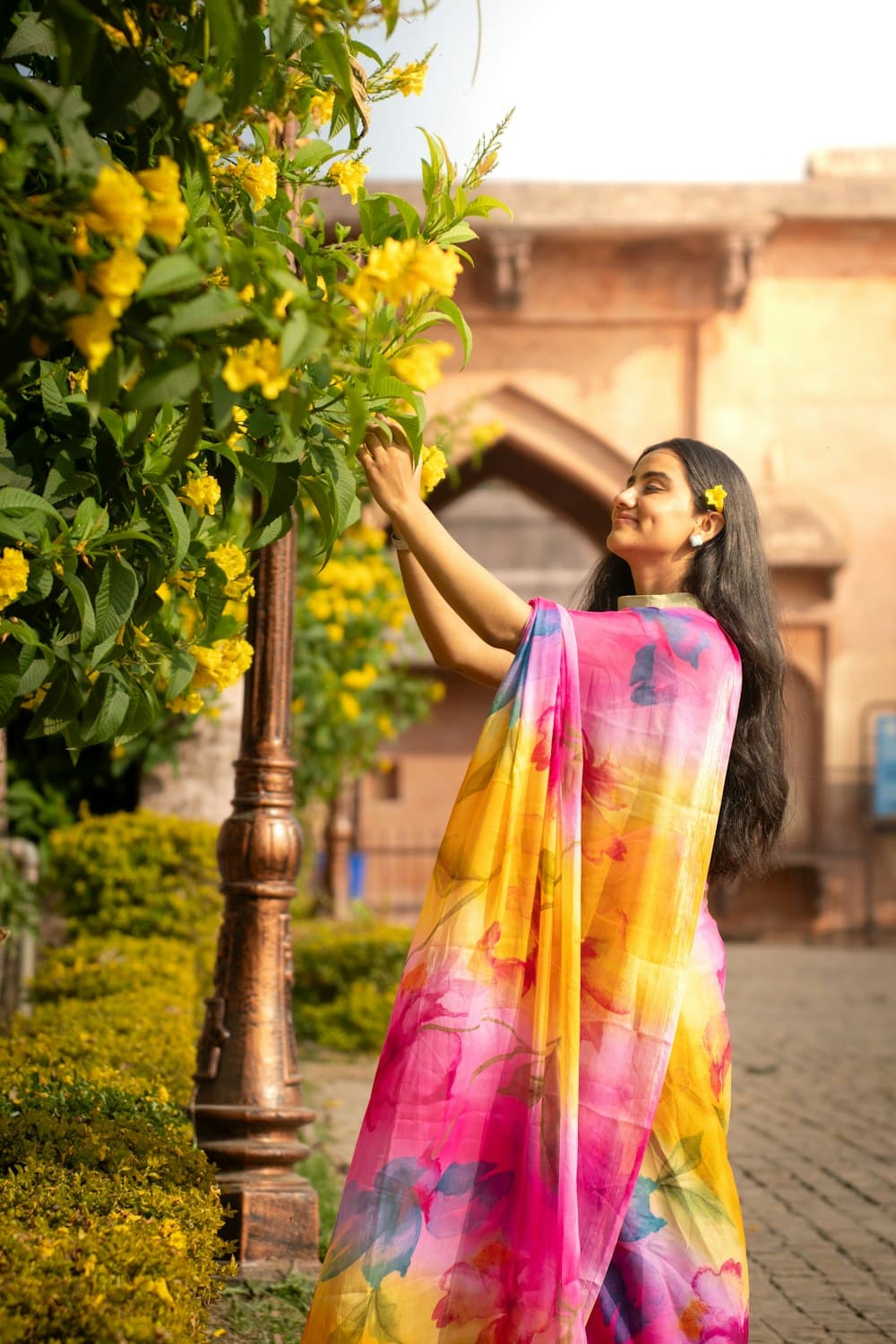 A woman in a bright floral saree reaches up towards yellow flowers