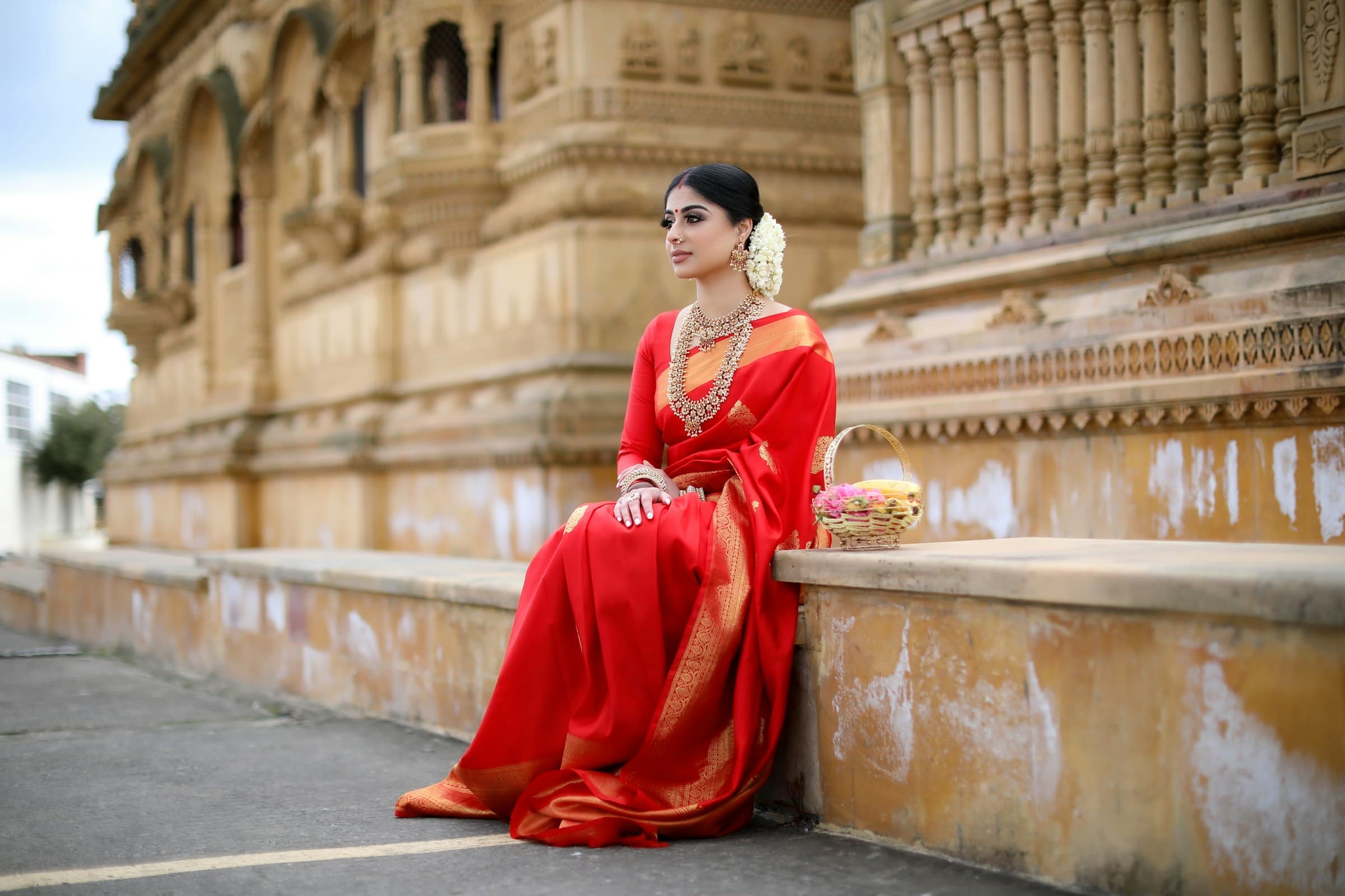 A woman in a red silk saree with a gold border sits on the steps of a carved sandstone temple